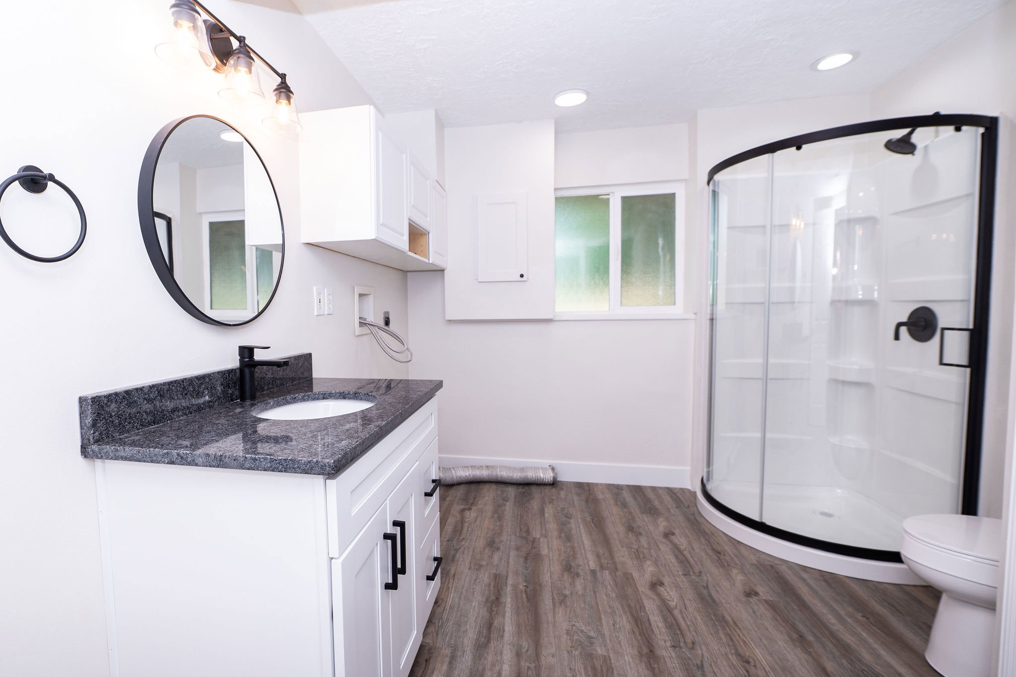 Bathroom featuring a curved glass shower enclosure, white vanity with a dark speckled countertop, black fixtures, and wood-finish flooring