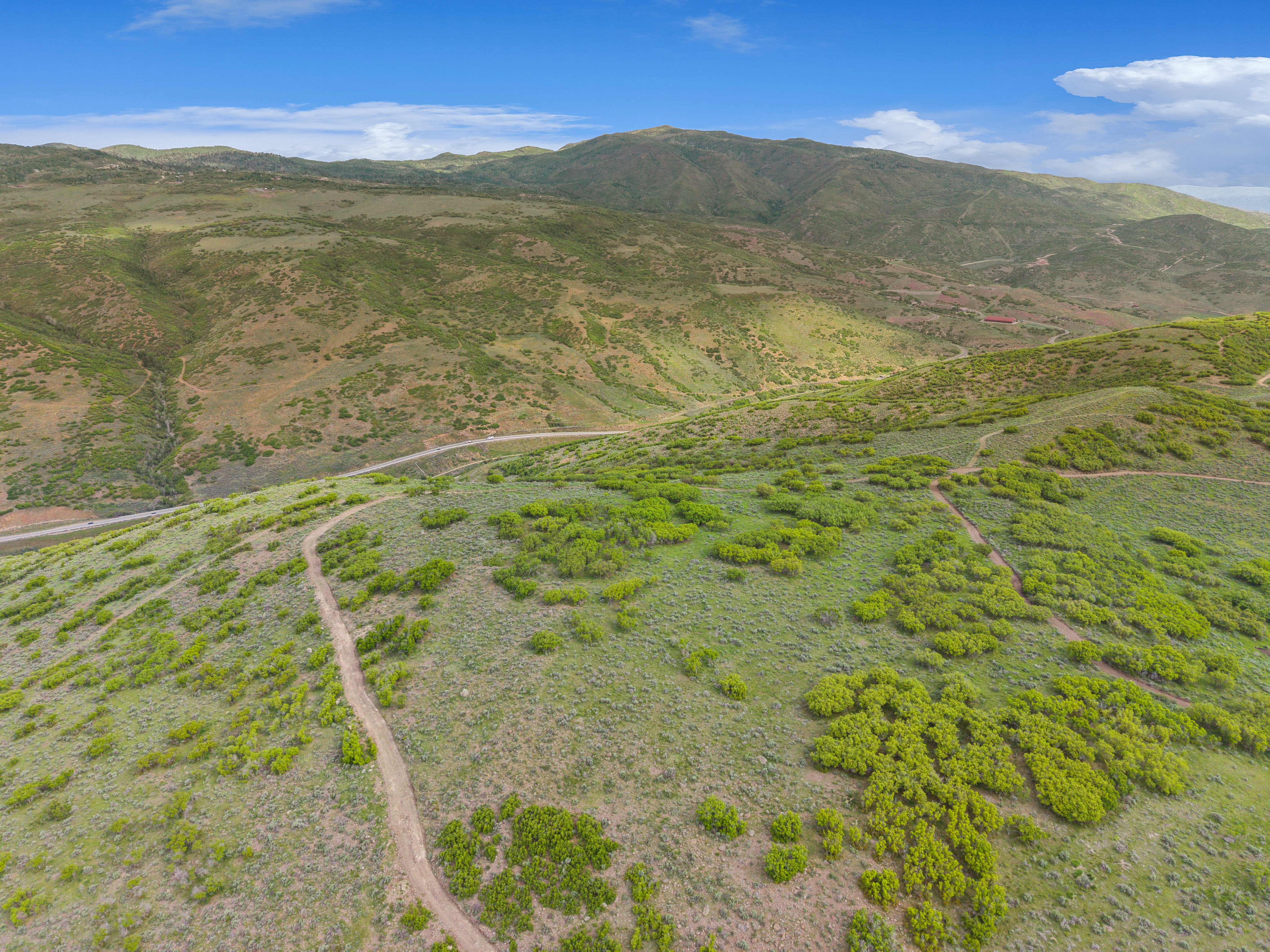 Expansive natural landscape featuring rolling hills, varied green vegetation, distant mountain ranges, and a winding dirt path