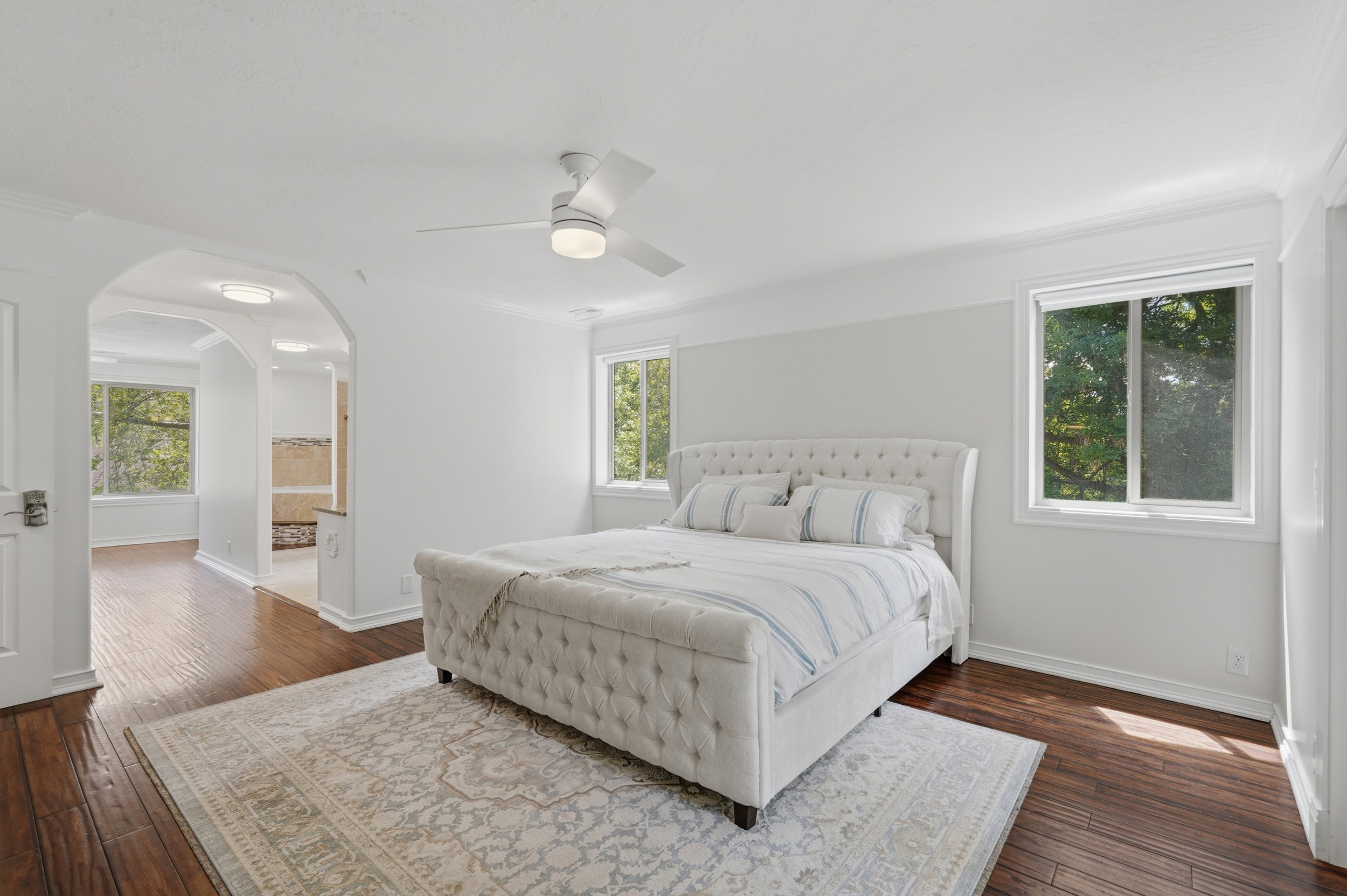 Bedroom with wood-finish flooring, two large windows, a white ceiling fan, and decorative archways leading to an additional space