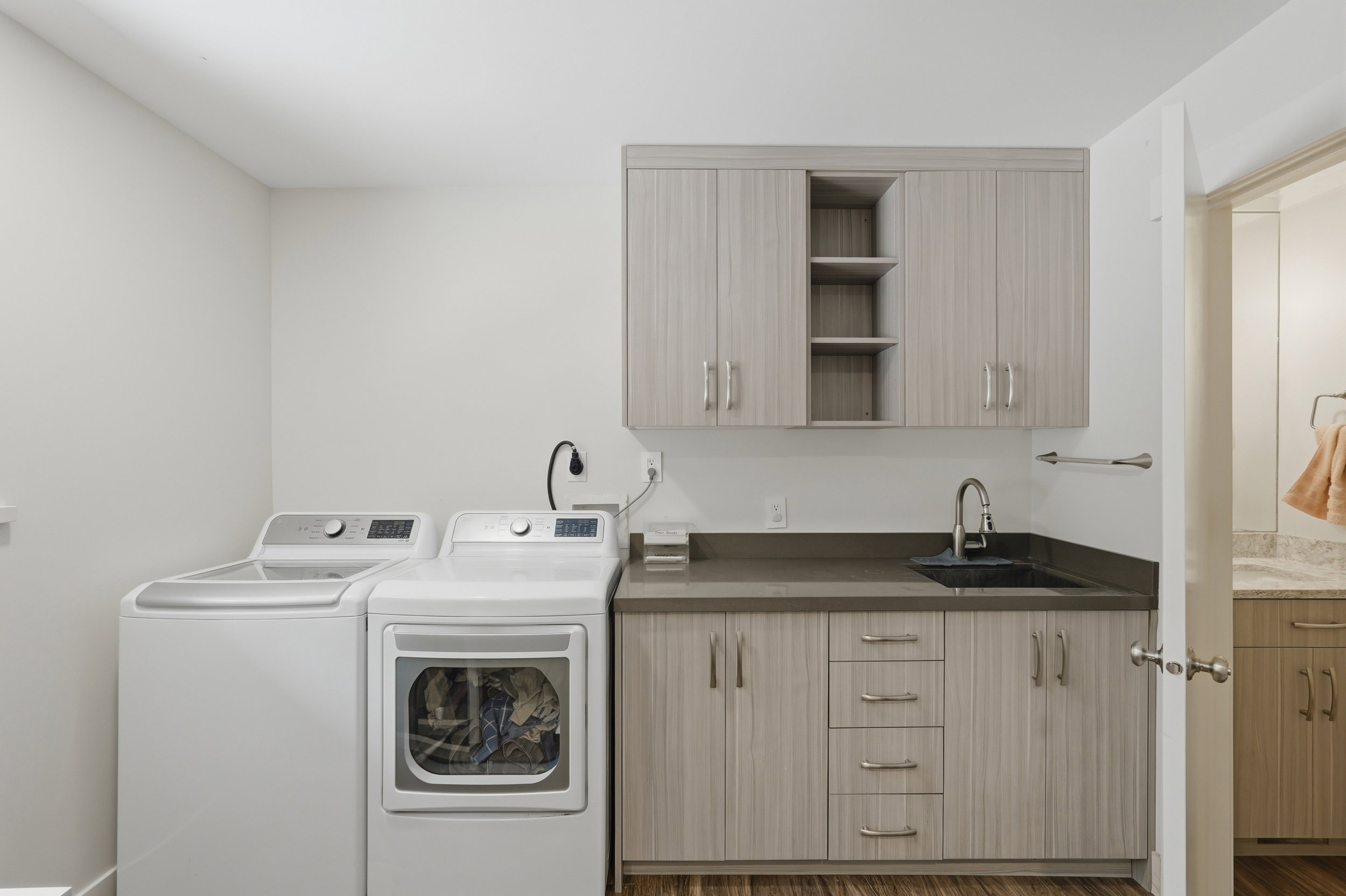 Dedicated laundry area featuring wood-finish flooring, light-toned cabinetry, a dark countertop, and a built-in sink with a gooseneck faucet