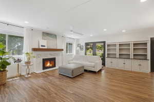 Spacious living area featuring wood-finish flooring, a fireplace with a white stone surround and wood mantel, recessed lighting, and built-in shelving with lower cabinetry