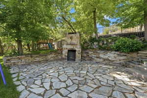 Stone flagstone patio with a built-in stone fireplace, featuring stacked stone retaining walls, mature trees, and a black wrought iron fence