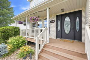 Front entryway featuring a covered porch with white railing, light brick siding, a dark entry door with decorative glass inserts, and landscaping with shrubs and rock ground cover