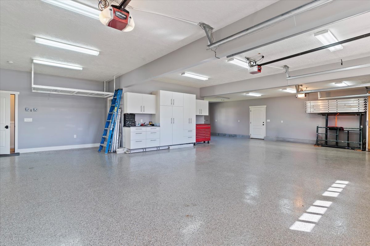 Spacious garage featuring a durable epoxy-coated concrete floor, extensive built-in white cabinetry, overhead storage racks, and multiple fluorescent lighting fixtures