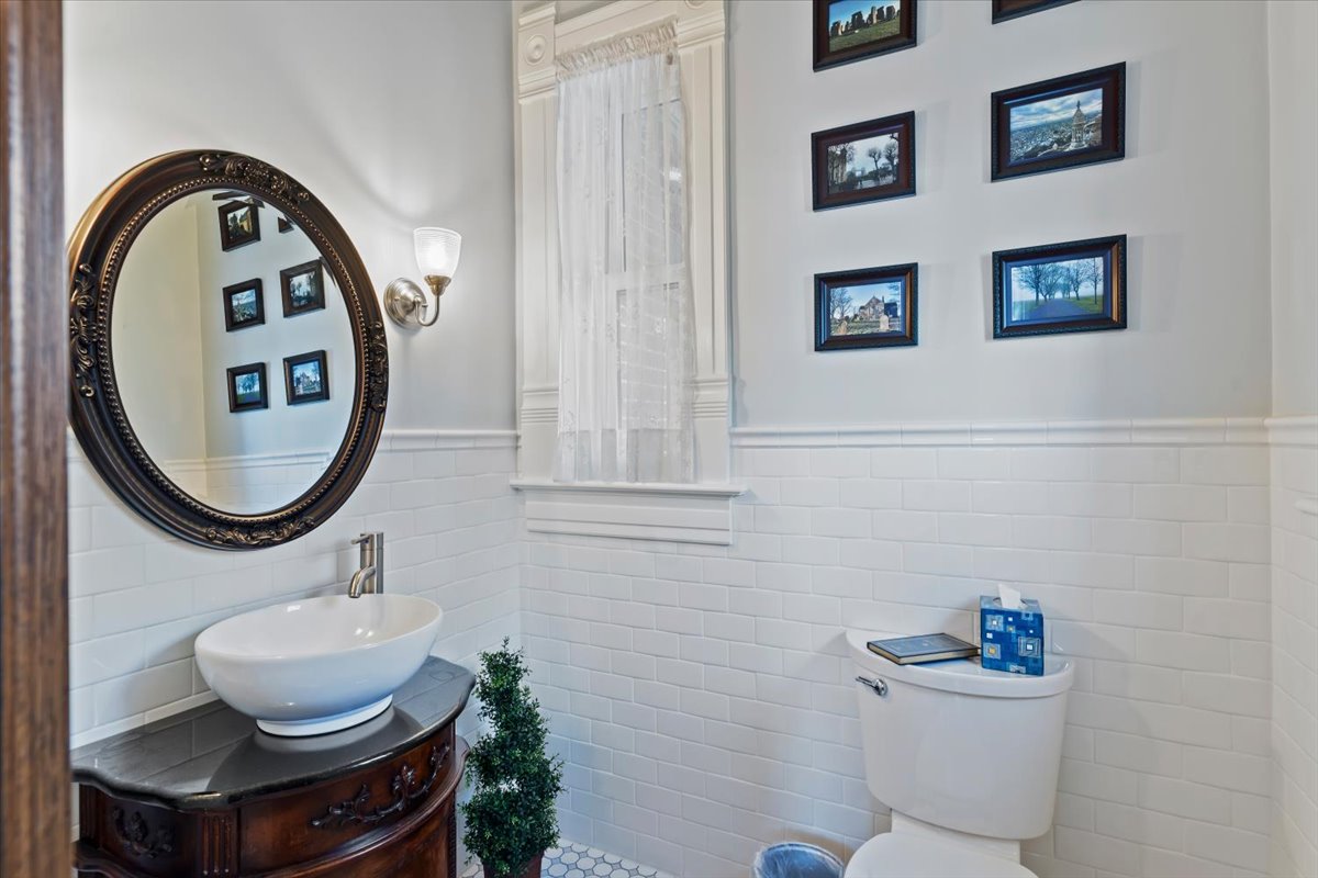 Bathroom featuring white subway tile wainscoting, an ornate oval mirror, a vessel sink with a dark wood vanity, a wall-mounted sconce, and white window trim