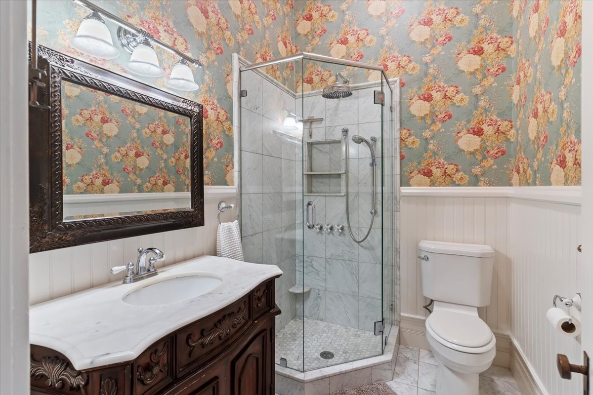 Bathroom featuring a glass-enclosed shower with tiled walls and floor, an ornate wood vanity with a white countertop and integrated sink, a dark framed mirror, and floral pattern wallpaper over white wainscoting