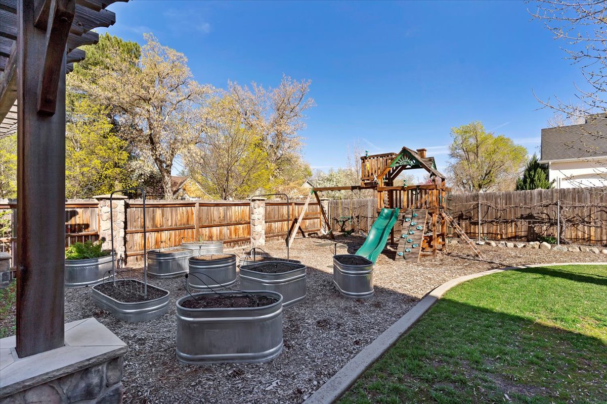 Backyard featuring a wood pergola structure, gravel ground cover, multiple metal raised garden beds, a wood privacy fence with stone pillars, and a manicured lawn area