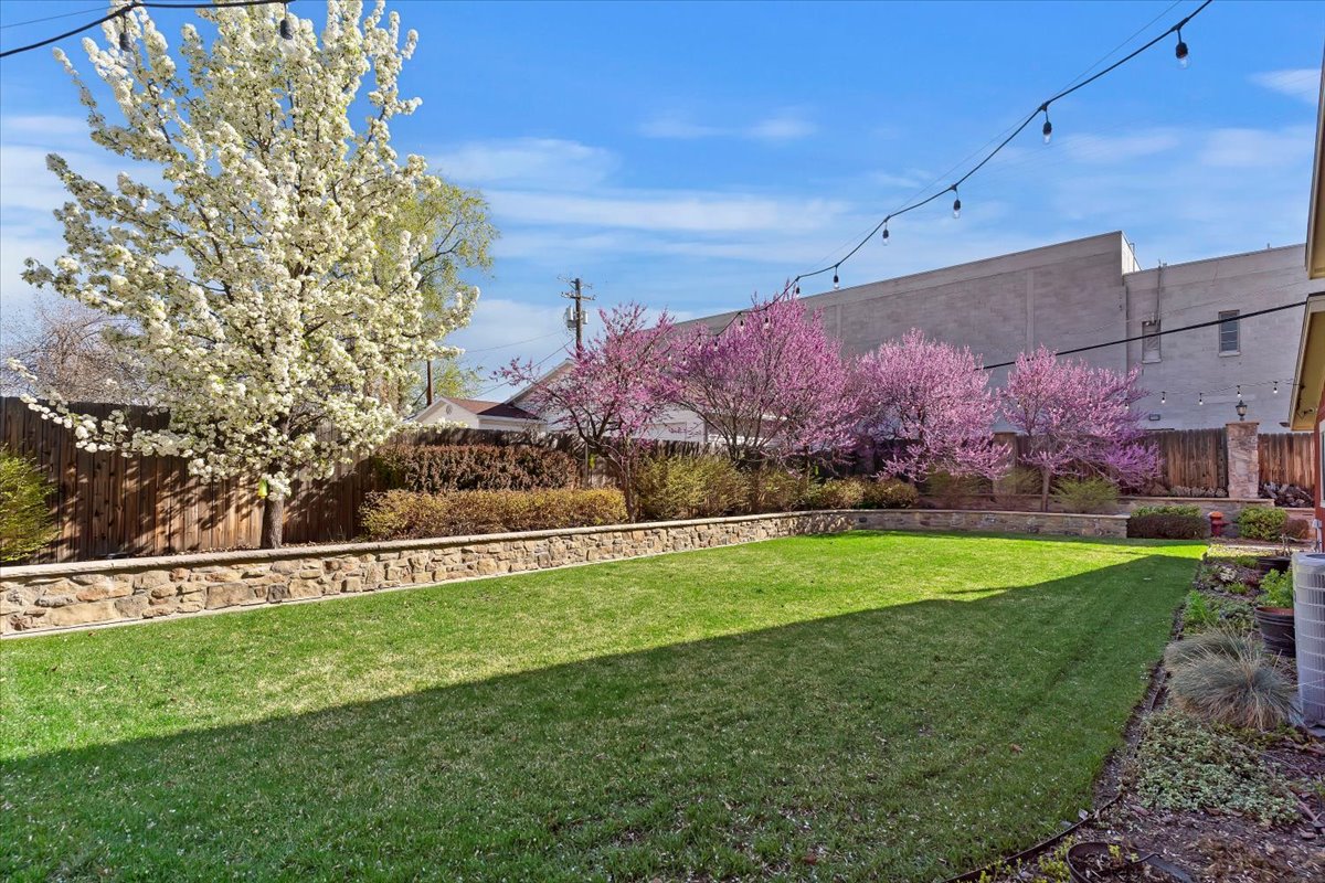 Expansive backyard featuring a manicured lawn, stone retaining walls, mature flowering trees, and a privacy fence