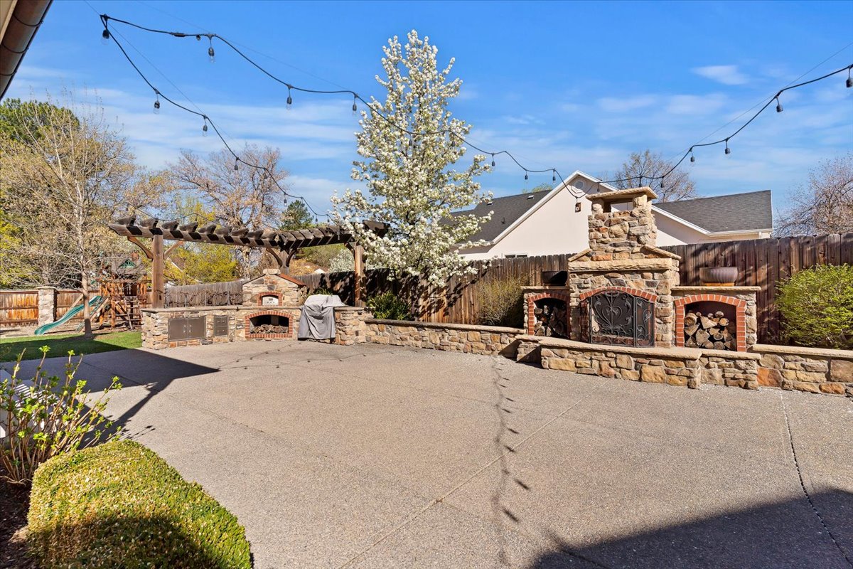 Expansive concrete patio area featuring a stone outdoor fireplace with integrated wood storage, a built-in outdoor kitchen with a pizza oven, and a wooden pergola structure