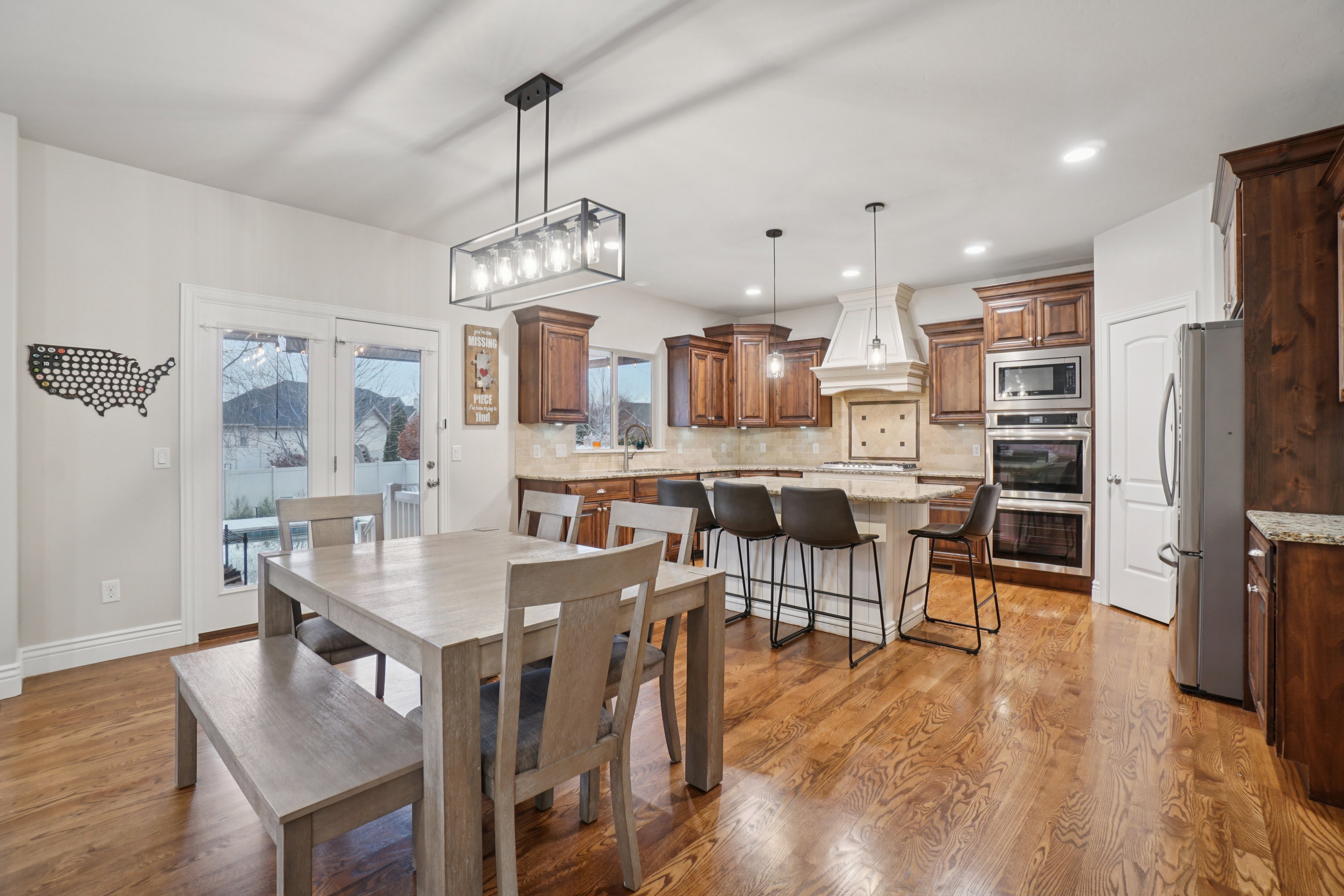 Open-concept kitchen and dining area featuring rich wood-finish flooring, a large kitchen island with seating, dark wood cabinetry, stainless steel double ovens, and a prominent range hood