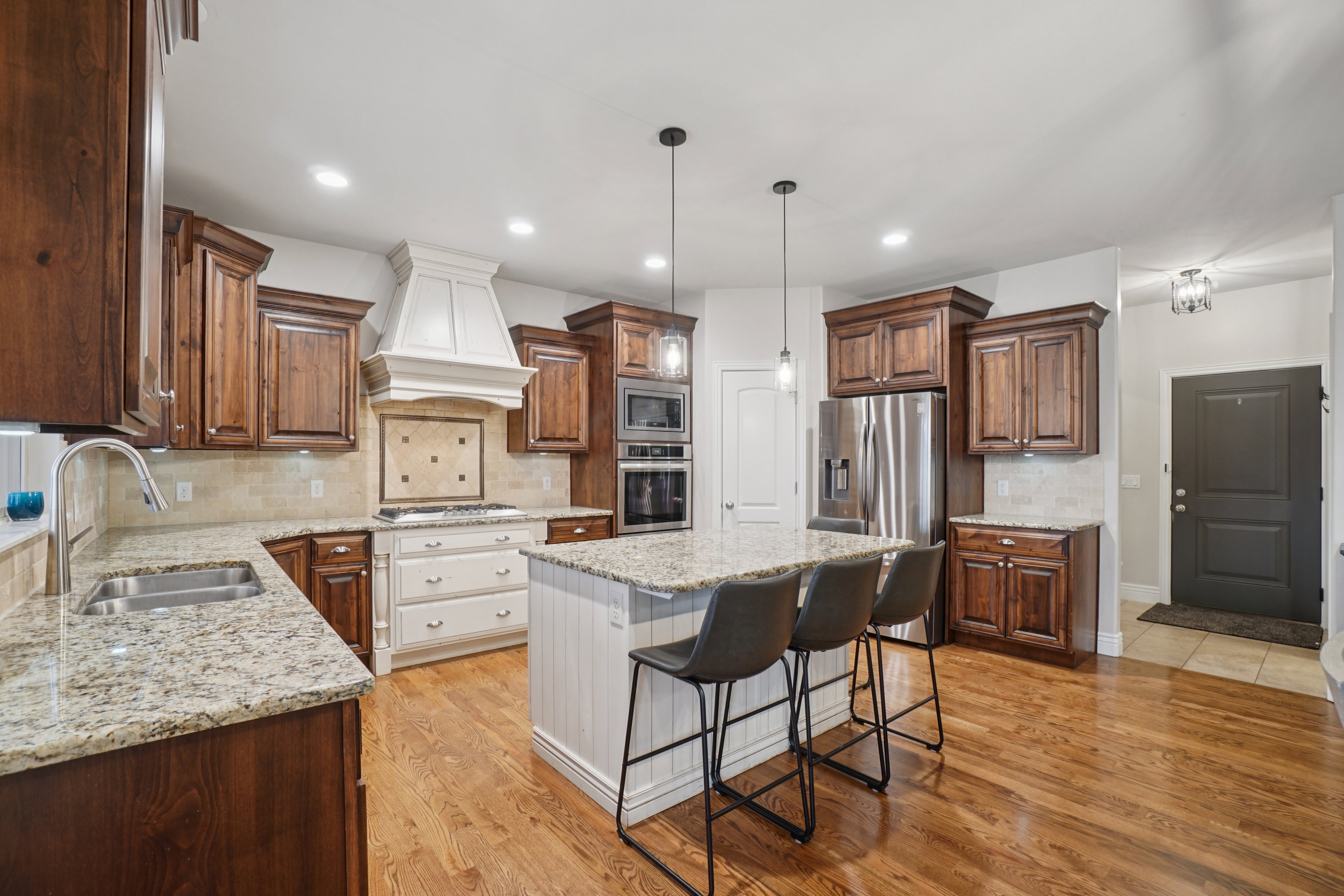 Kitchen featuring wood-finish flooring, granite countertops, stainless steel appliances, a center island with pendant lighting, and a white range hood with a tiled backsplash