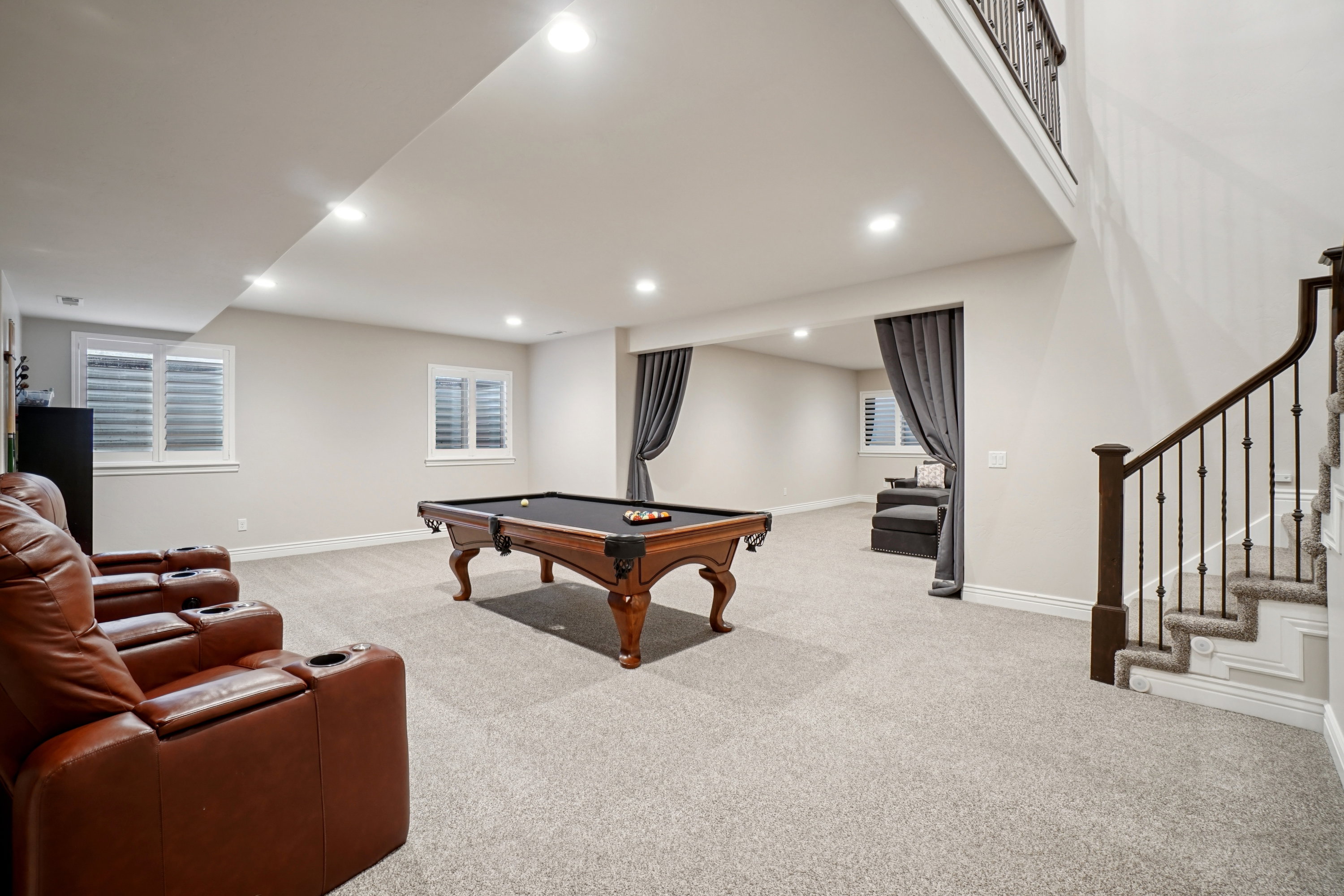 Expansive recreation room featuring recessed lighting, light-colored carpet, multiple windows with white plantation shutters, and an open staircase with dark wood newel posts and iron balusters
