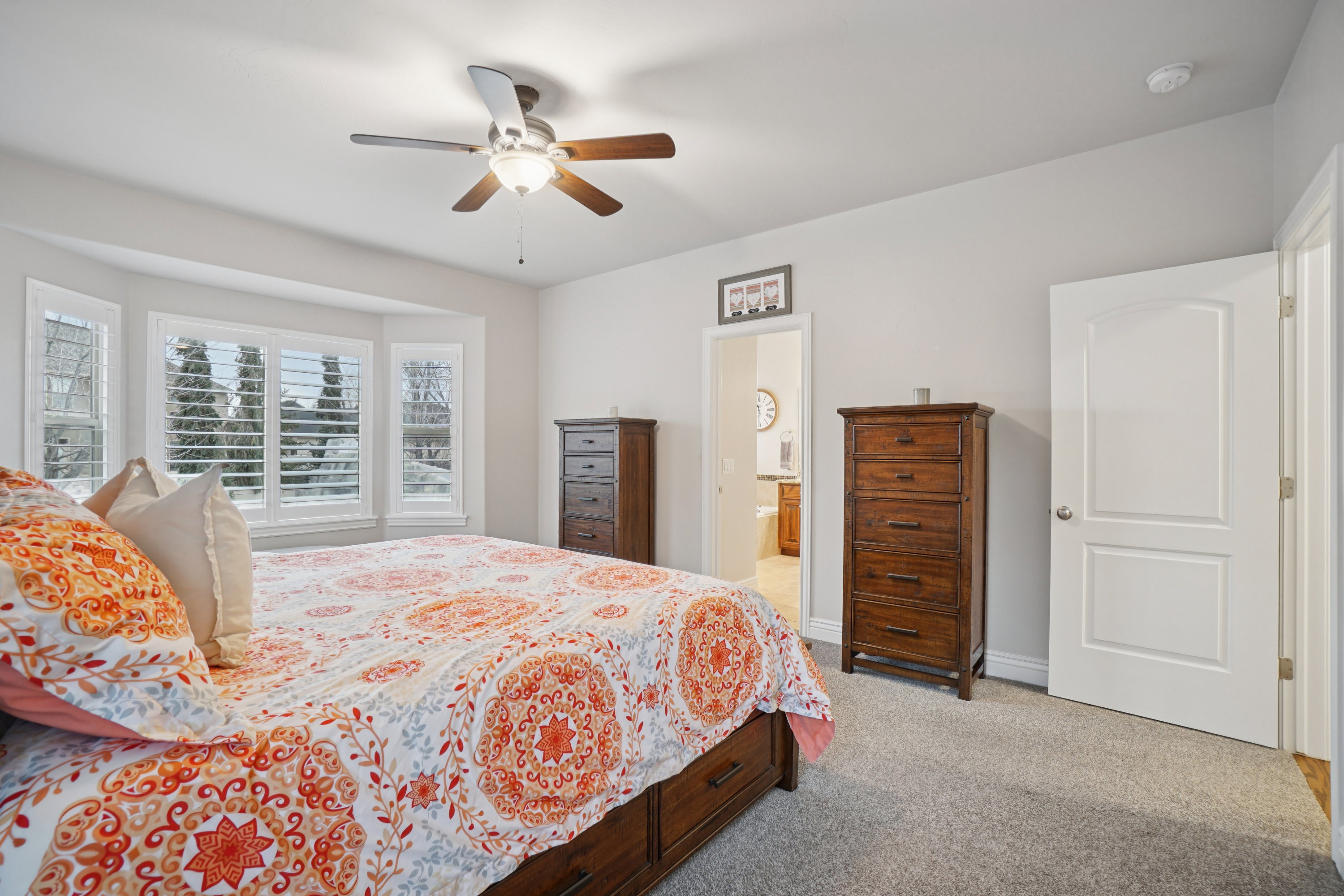 Spacious room featuring a bay window with plantation shutters, soft gray carpeting, and an overhead ceiling fan with lighting