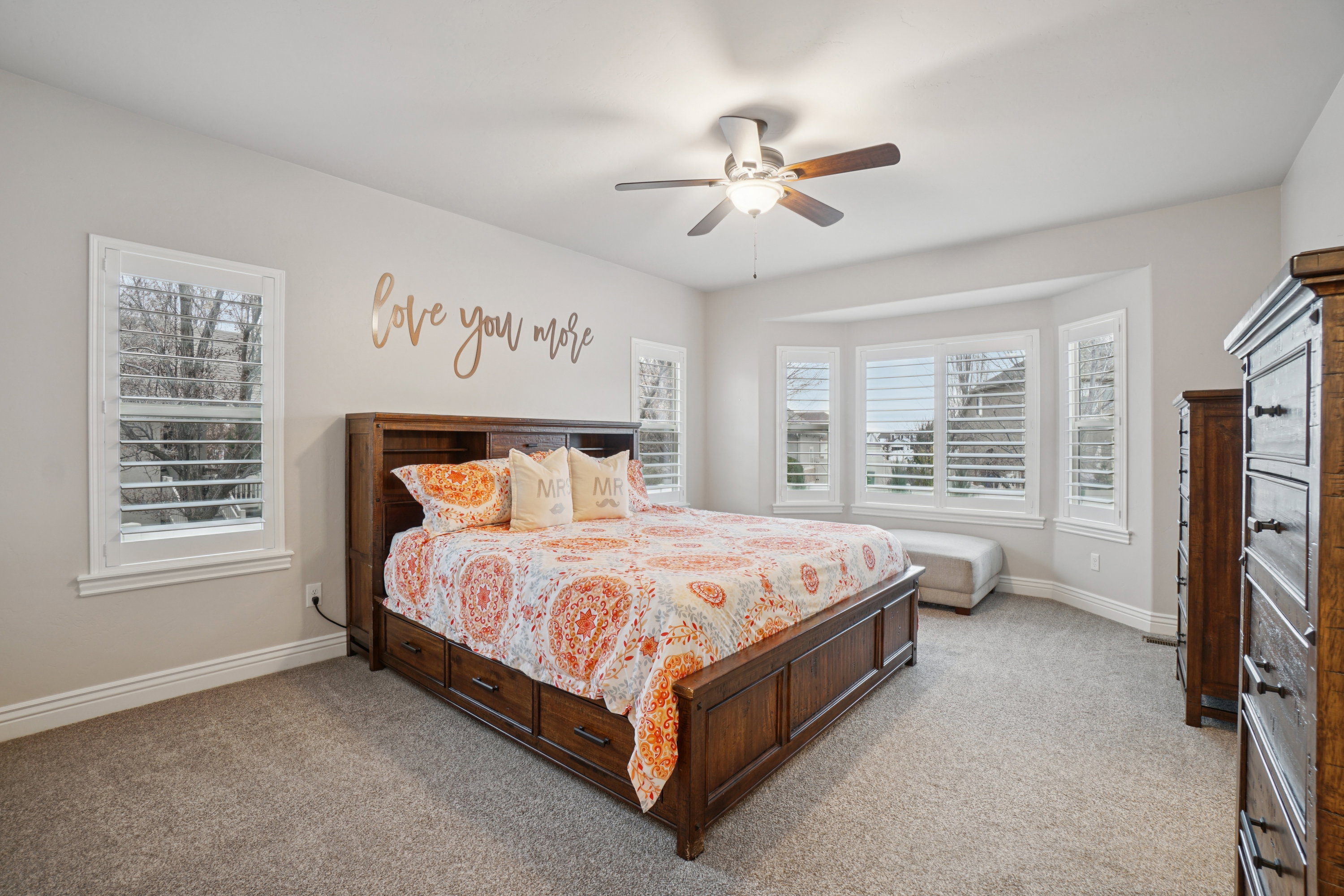 Spacious room featuring a bay window with plantation shutters, additional window with plantation shutters, ceiling fan, and neutral carpeting