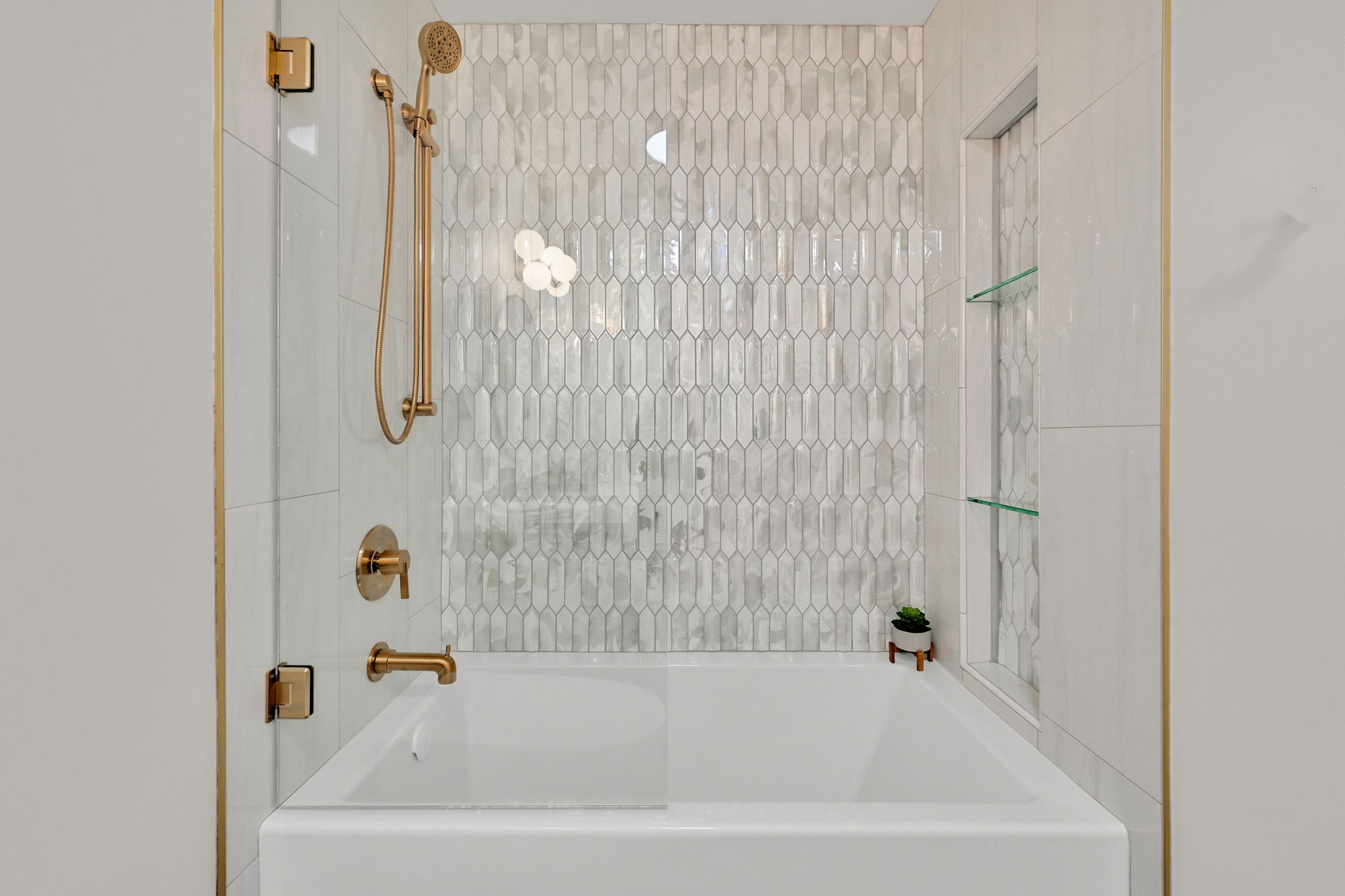 Bathtub with a unique picket tile surround, brushed gold fixtures, a built-in shower niche with glass shelving, and a clear glass shower panel