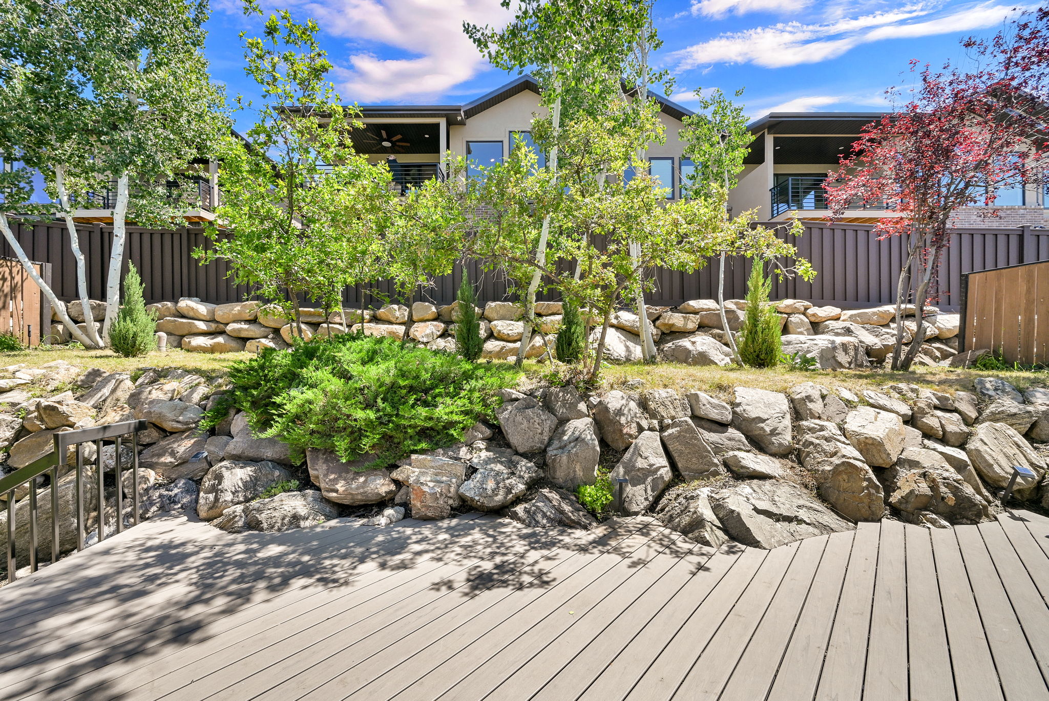 Composite decking with dark railing, backed by a rock retaining wall and slatted fencing