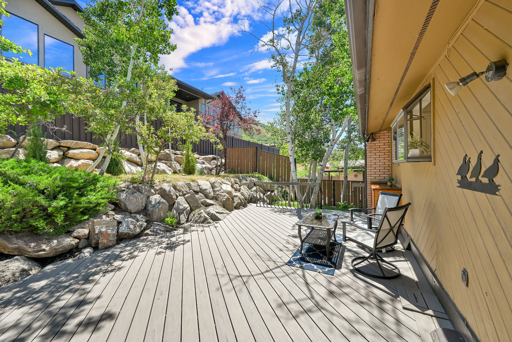 Expansive outdoor deck featuring composite decking, a rock retaining wall with established landscaping, and mature trees