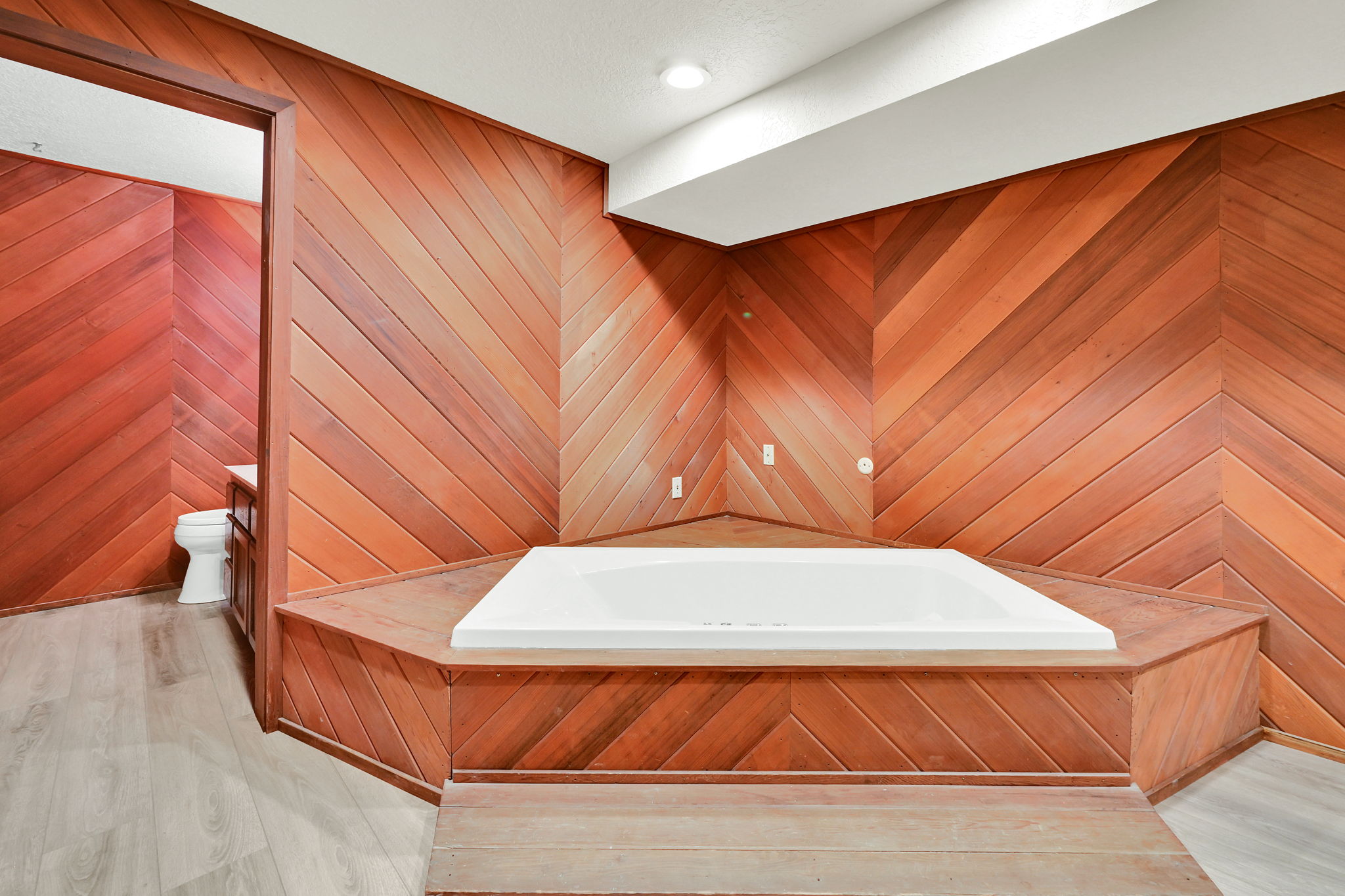 Built-in corner soaking tub featuring wood-finish paneling, chevron-patterned wood-finish walls, and light wood-finish flooring