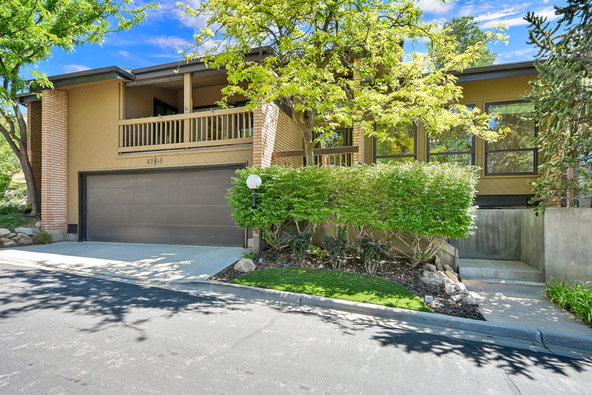 Contemporary exterior with a two-car garage, upper-level balcony, brick accents, and mature landscaping