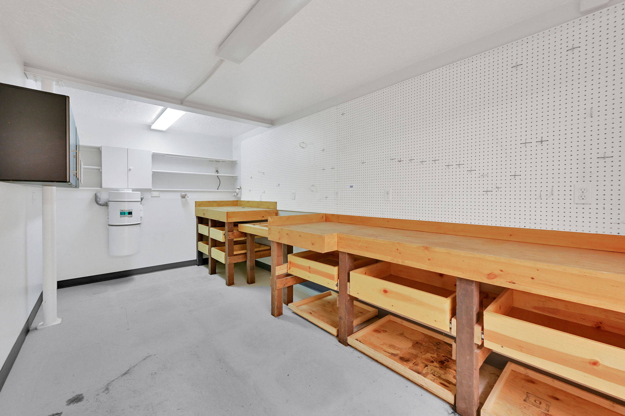 Dedicated workshop space featuring a white pegboard wall, multiple built-in wood workbenches with integrated storage, overhead fluorescent lighting, and white cabinetry