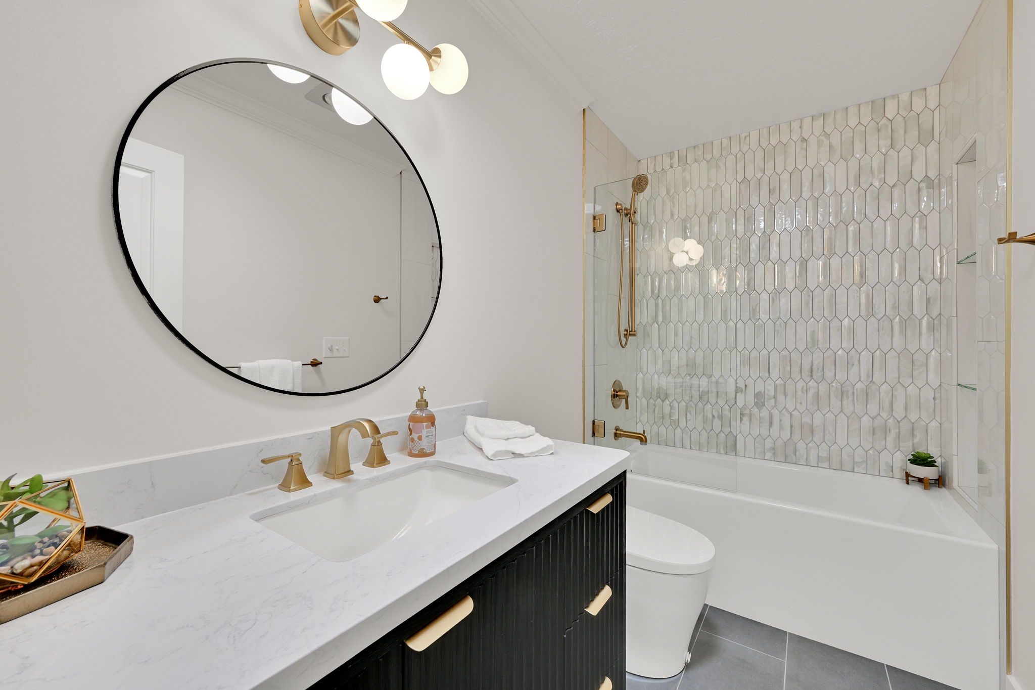 Bathroom featuring a contemporary vanity with a white countertop, gold-toned fixtures, a circular mirror, and a tiled shower-tub combination with a glass enclosure