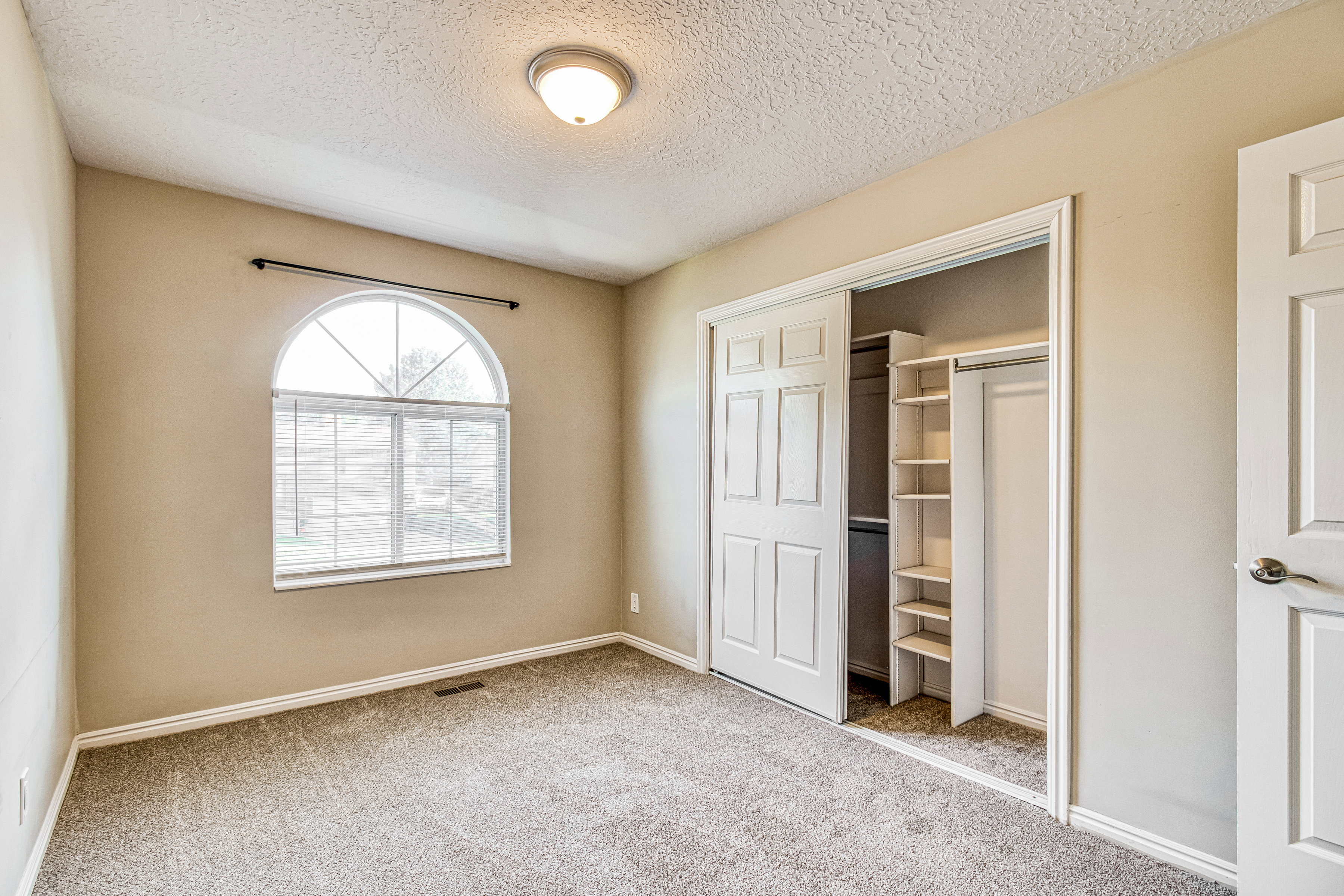 Room featuring a large arched window with horizontal blinds, neutral carpet flooring, and a built-in closet system with shelving and hanging space