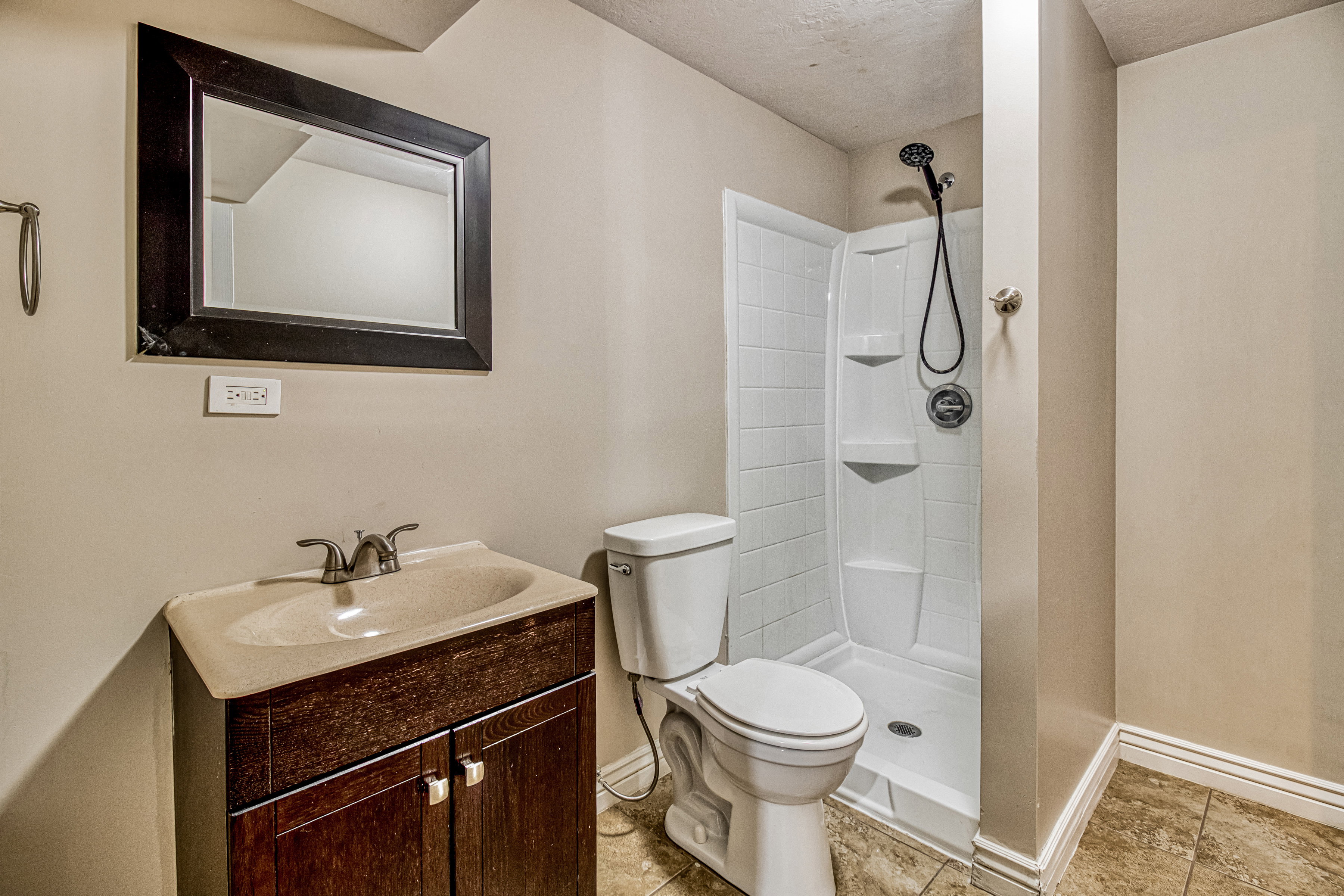Bathroom featuring a dark wood-finish vanity with an integrated sink, a framed mirror, a standalone shower with a handheld fixture, and tile flooring