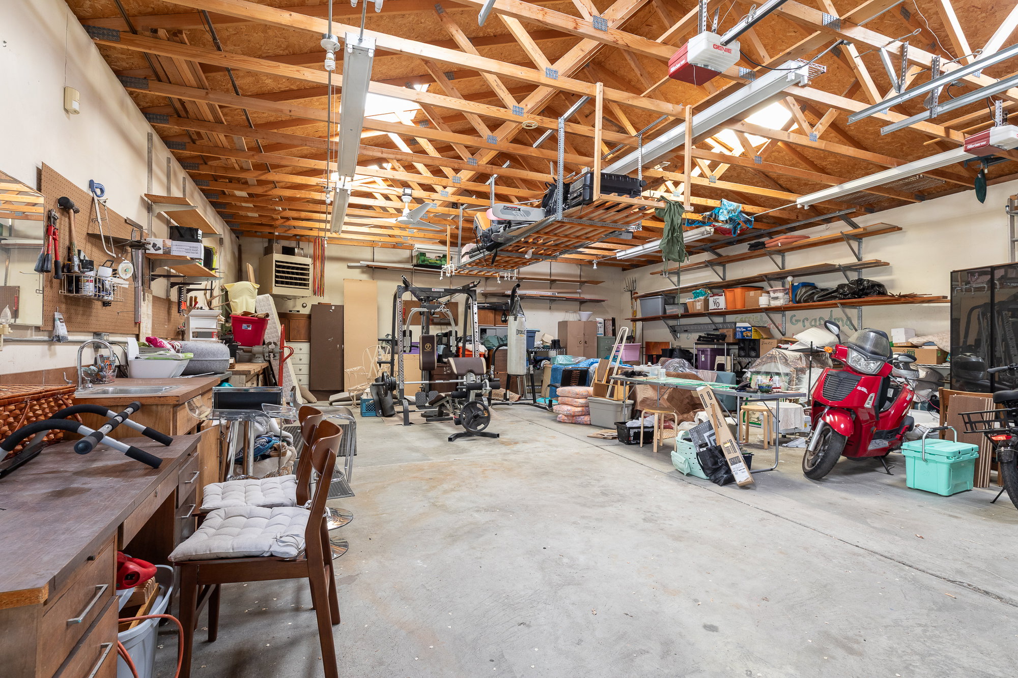 Spacious garage featuring exposed wood beam ceilings, concrete flooring, ample built-in shelving, a workbench with an integrated sink, and overhead storage racks