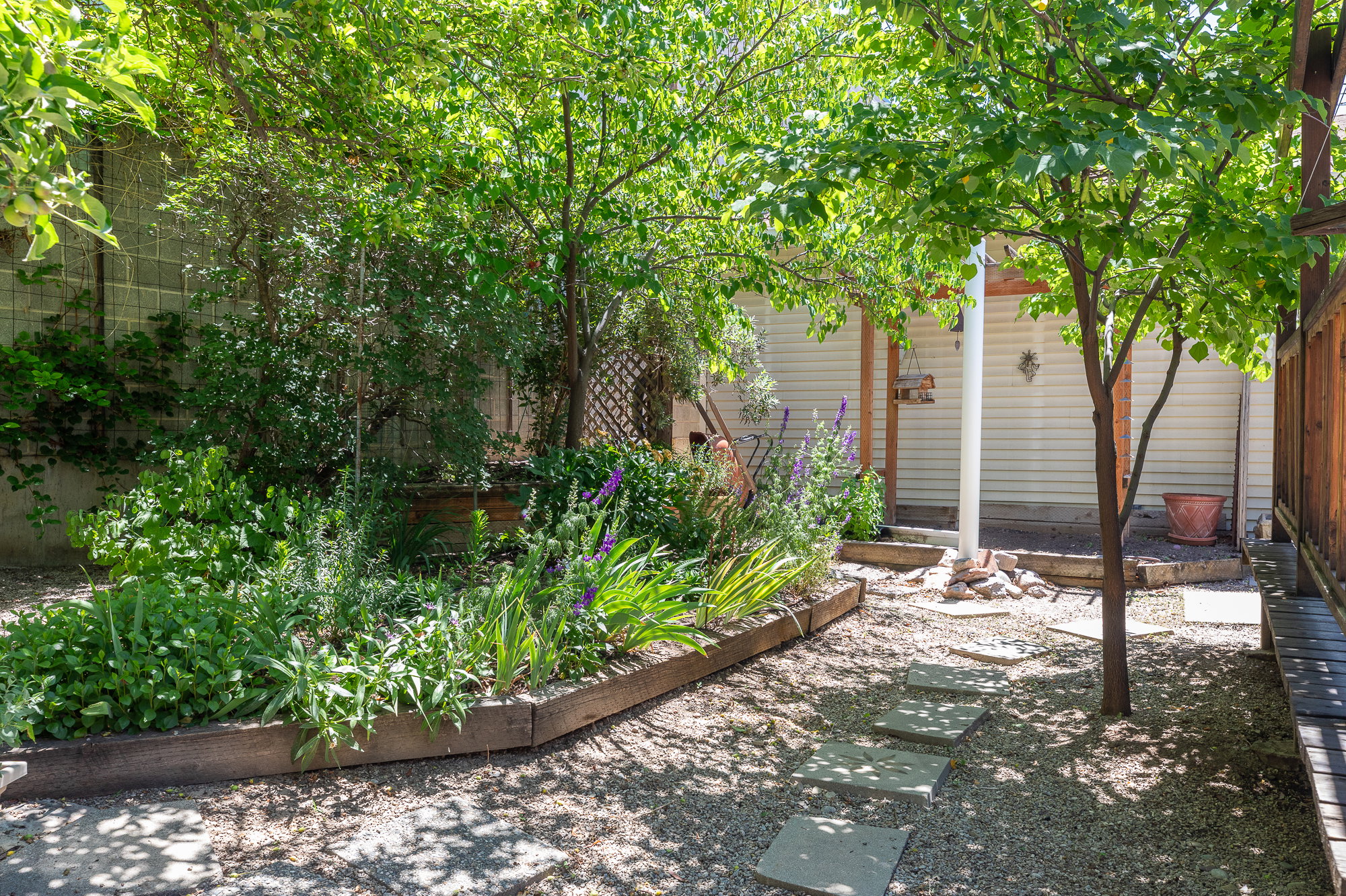 Gravel patio area featuring a stone paver pathway, mature shade trees, raised garden beds with established plantings, and a wood-slat fence