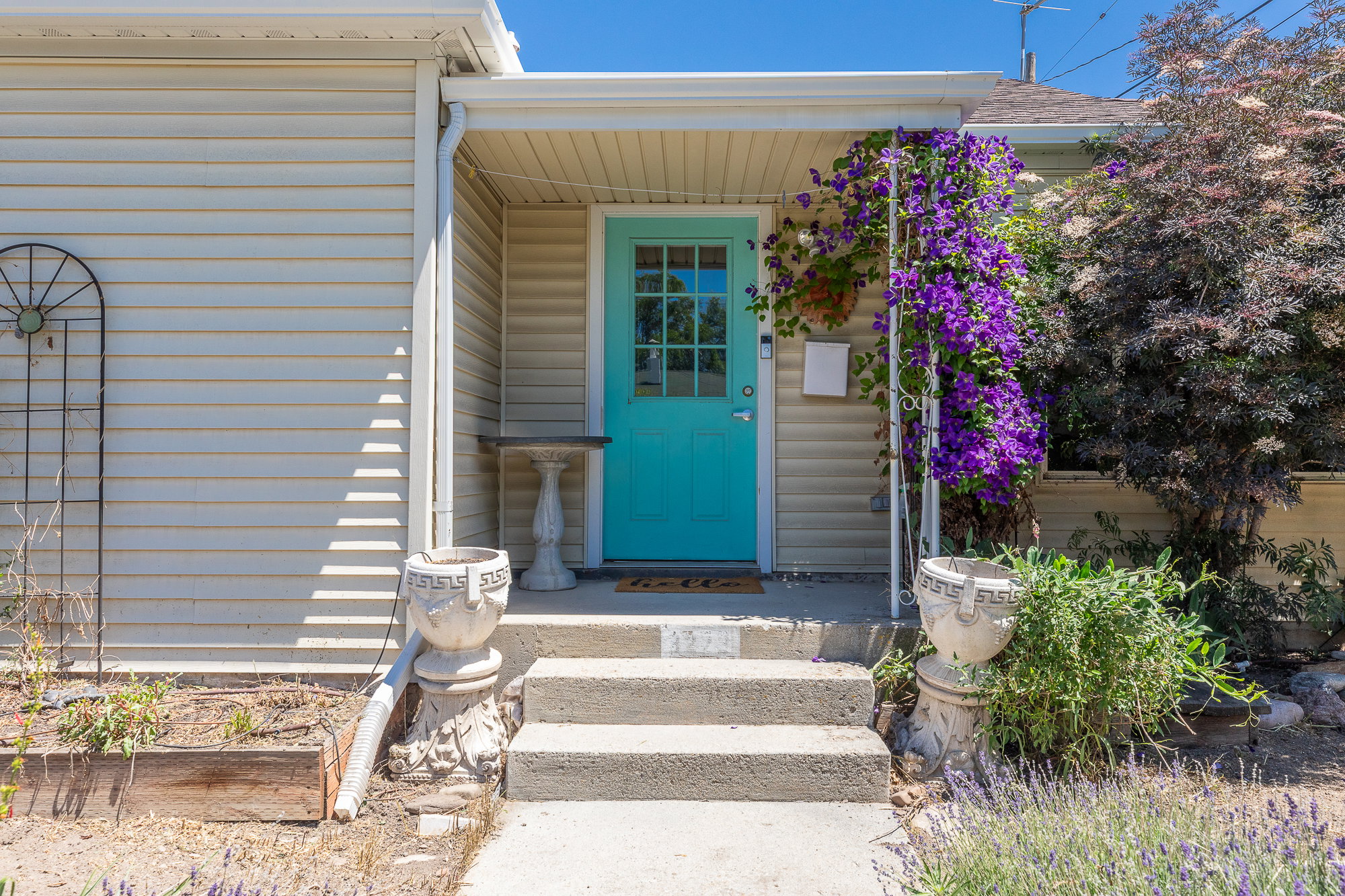 Light-colored horizontal siding exterior featuring a bright turquoise door with divided lites, a covered porch, and mature purple flowering vines
