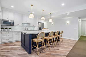 Gourmet kitchen featuring a large central island with seating, white shaker cabinetry, stainless steel appliances, and wood-finish flooring
