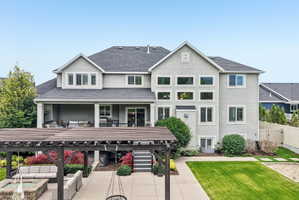Rear exterior featuring a large stucco facade, multiple window configurations, a covered patio with white columns, a dark wood pergola over a paved patio, and a manicured lawn