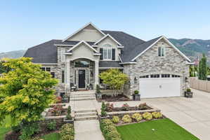 Stately stone and stucco exterior featuring a prominent arched entryway, a paved driveway, an attached two-car garage, mature landscaping, and a mountain backdrop