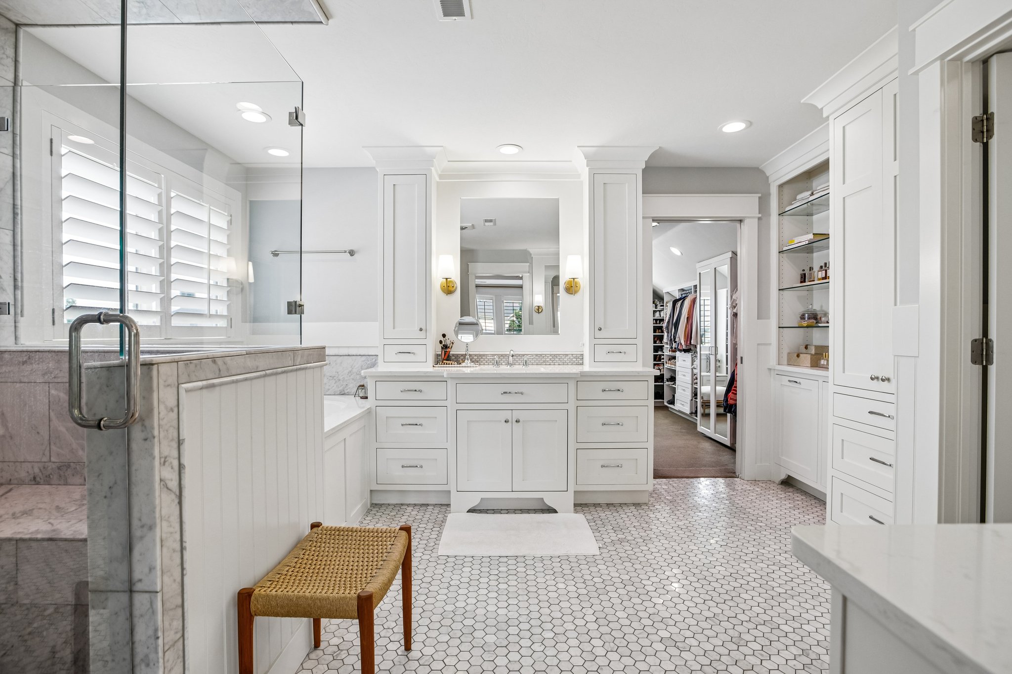 Spacious bathroom featuring white hexagonal tile flooring, a glass-enclosed shower, a built-in soaking tub, a double vanity with stone countertops, and extensive built-in cabinetry with open shelving
