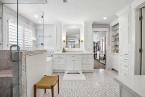 Spacious bathroom featuring white hexagonal tile flooring, a glass-enclosed shower, a built-in soaking tub, a double vanity with stone countertops, and extensive built-in cabinetry with open shelving