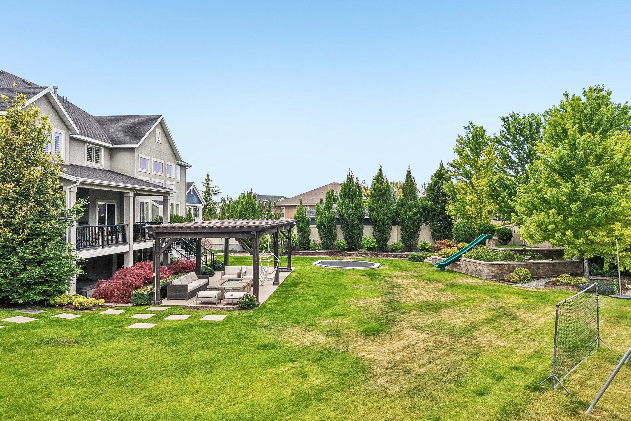 Expansive backyard featuring a large lawn area, a paved patio with a wooden pergola, a raised garden bed with a slide, and mature trees providing natural screening