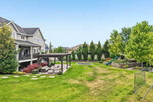 Expansive backyard featuring a large lawn area, a paved patio with a wooden pergola, a raised garden bed with a slide, and mature trees providing natural screening