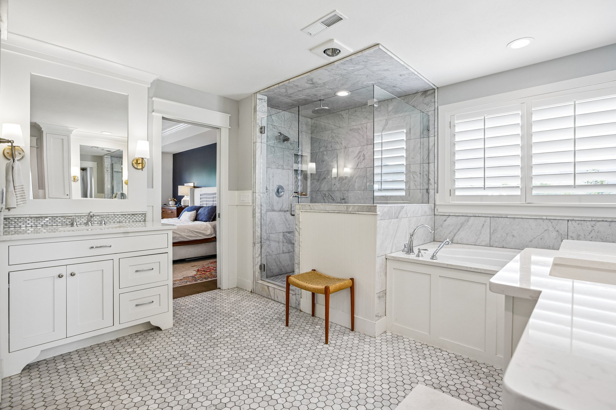 Bathroom featuring white hexagon tile flooring, a glass-enclosed shower with tiled walls, a separate soaking tub with a tiled surround, white vanity cabinetry with a stone countertop, and plantation shutters