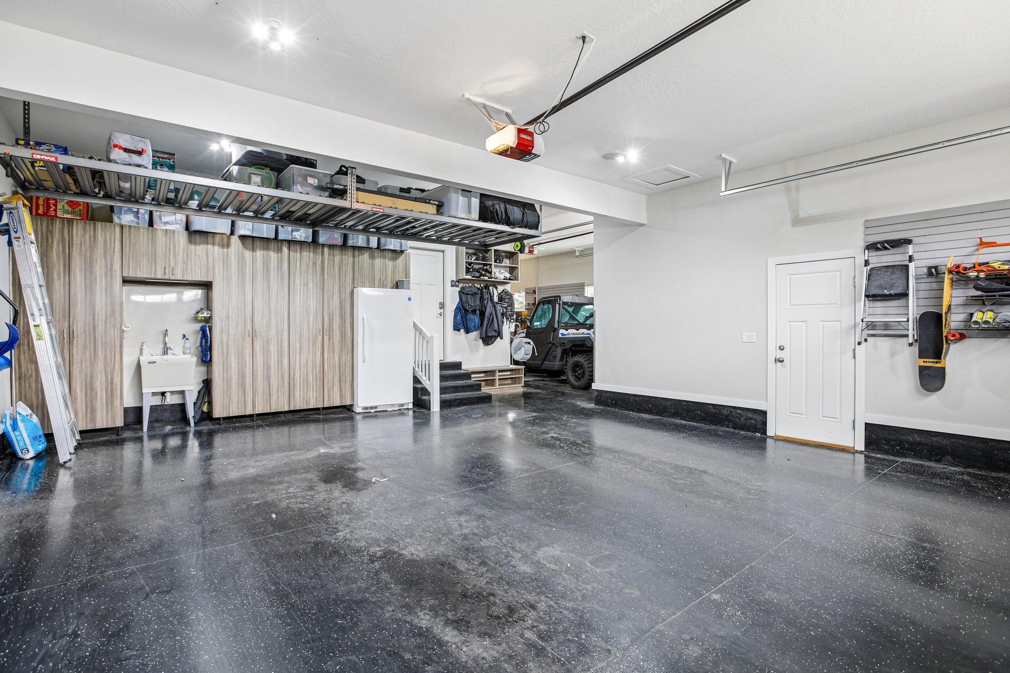 Spacious garage featuring a durable dark flooring finish, extensive built-in wood-finish cabinetry, an integrated utility sink, overhead storage racks, and a white entry door