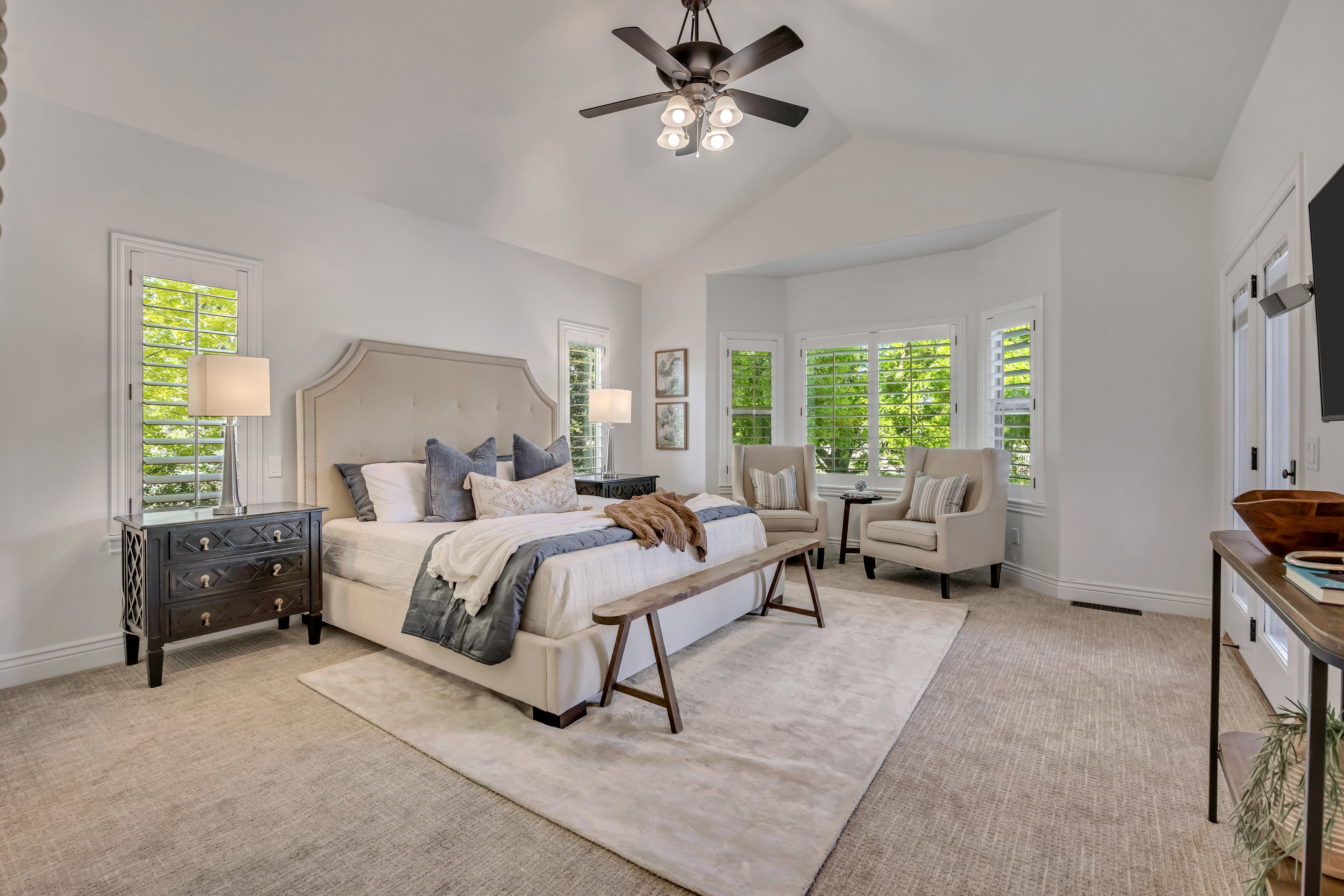 Vaulted ceiling with a ceiling fan, bay window seating area, plantation shutters, neutral-toned carpeting, and an upholstered bed frame