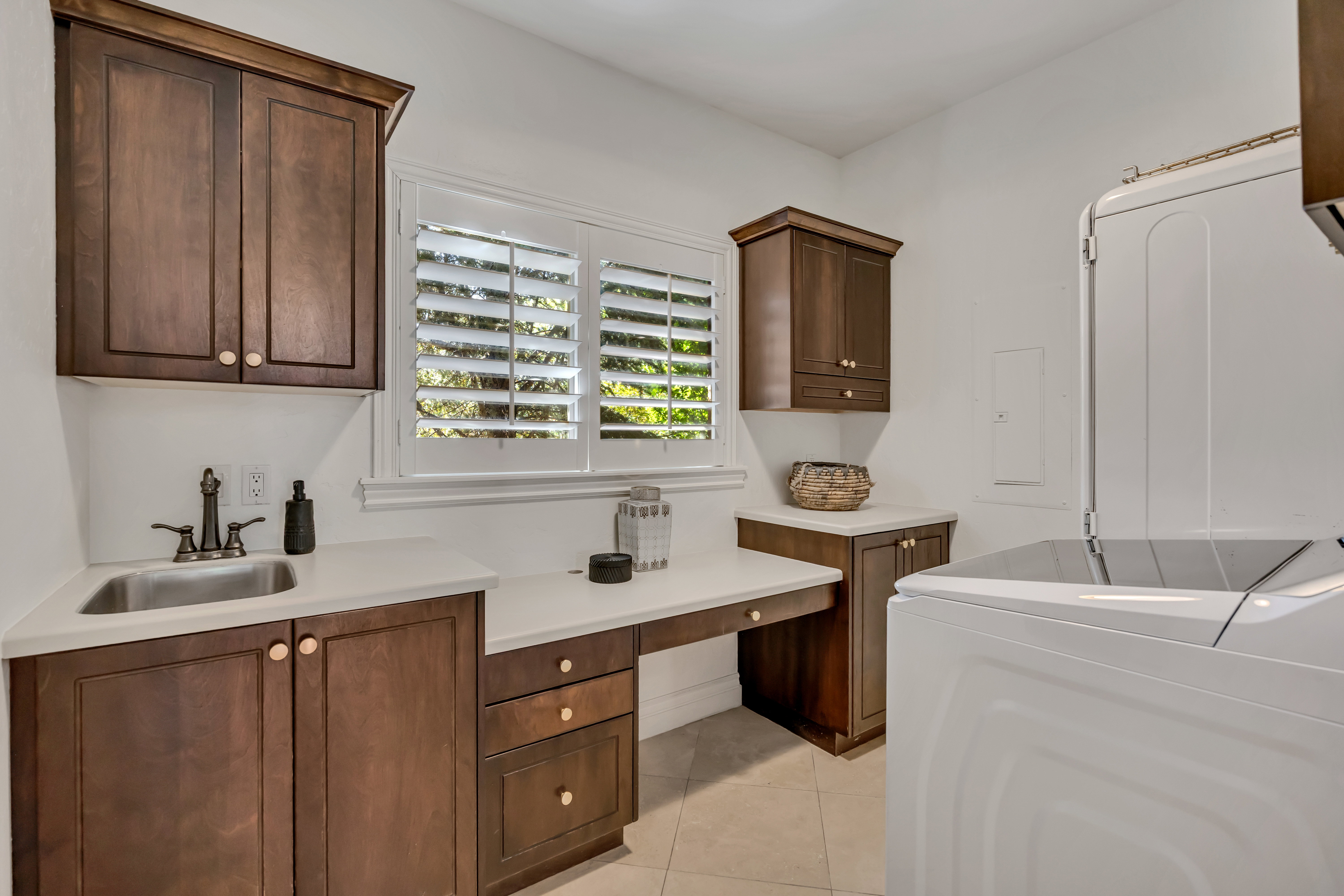 Laundry room featuring white shaker shutters, extensive wood cabinetry, a built-in desk area, a stainless steel utility sink, and light-toned tile flooring