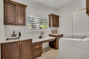Laundry room featuring white shaker shutters, extensive wood cabinetry, a built-in desk area, a stainless steel utility sink, and light-toned tile flooring