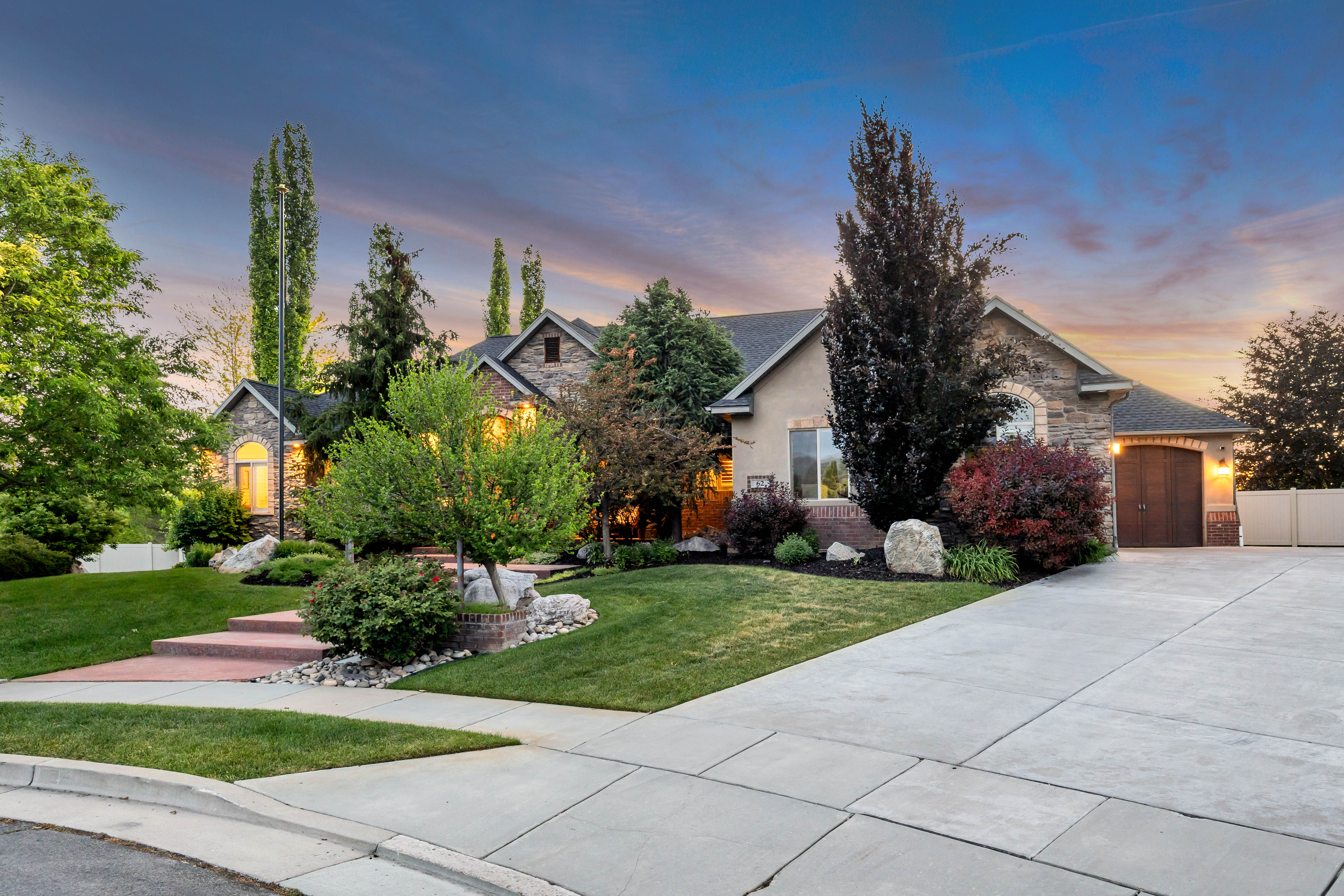 Stone and stucco exterior featuring a gabled roofline, mature landscaping, a concrete driveway, and an arched garage door
