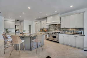 Kitchen featuring a substantial central island with integrated sink, stainless steel range with custom vent hood, light-toned cabinetry, natural stone countertops, and tile flooring