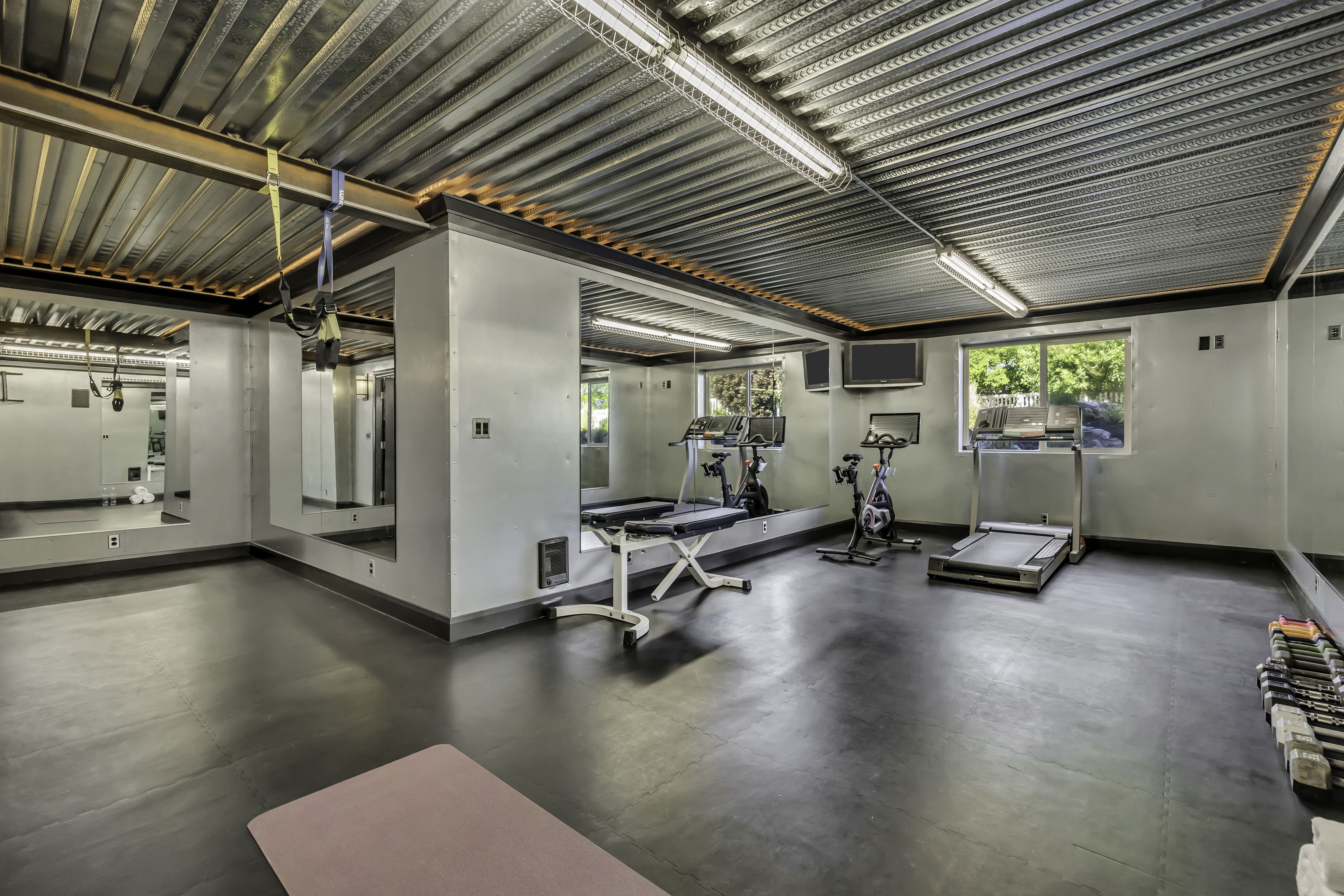 Dedicated fitness room featuring dark rubber flooring, mirrored walls, a corrugated metal ceiling with exposed beams, and fluorescent strip lighting