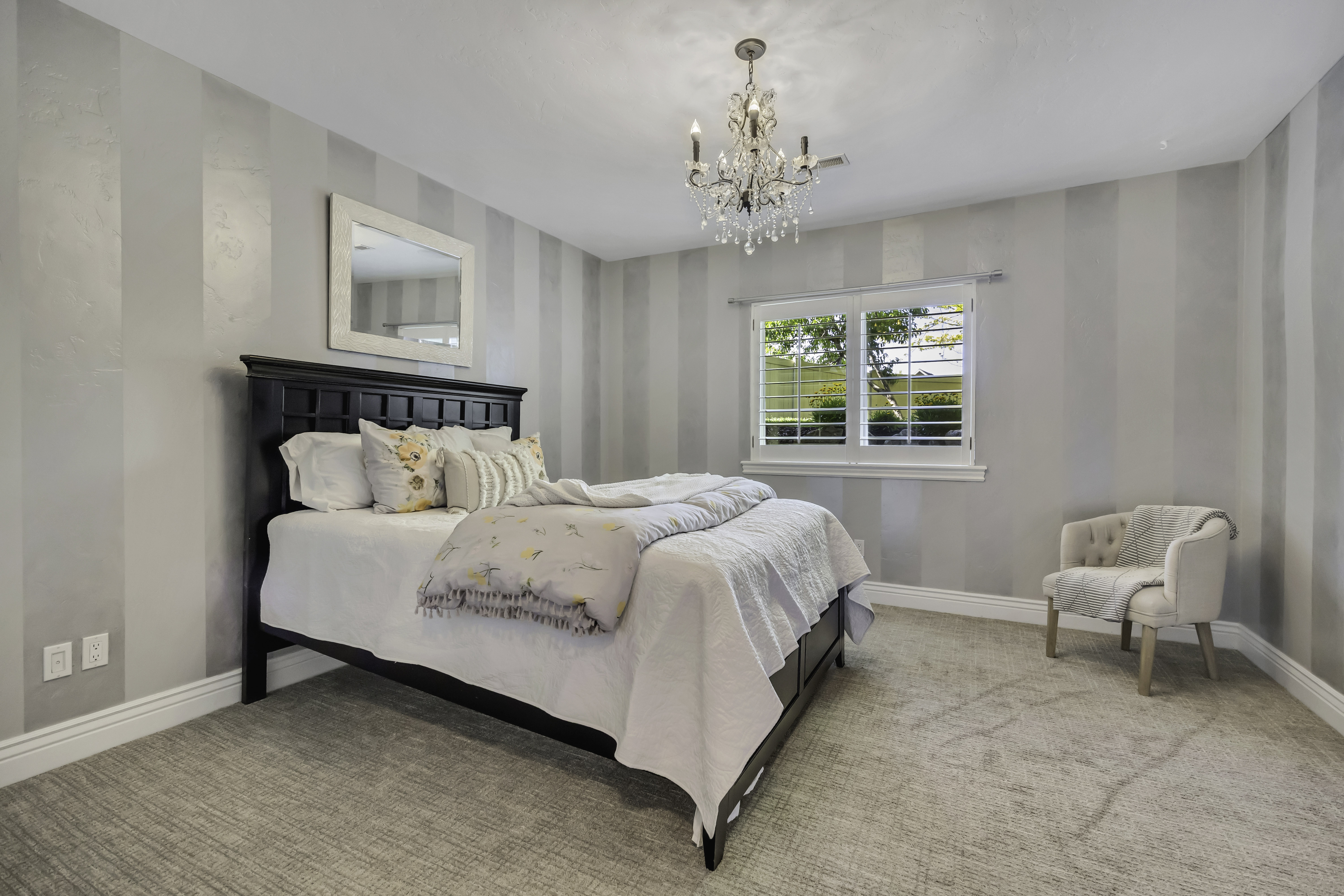 Bedroom featuring striped wall treatment, ceiling-mounted chandelier, and window with plantation shutters