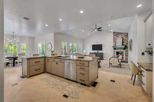 Open-concept living space featuring a kitchen island with stone countertops and integrated sink, stainless steel dishwasher, vaulted ceiling with recessed lighting, and a stone fireplace with a wood mantel