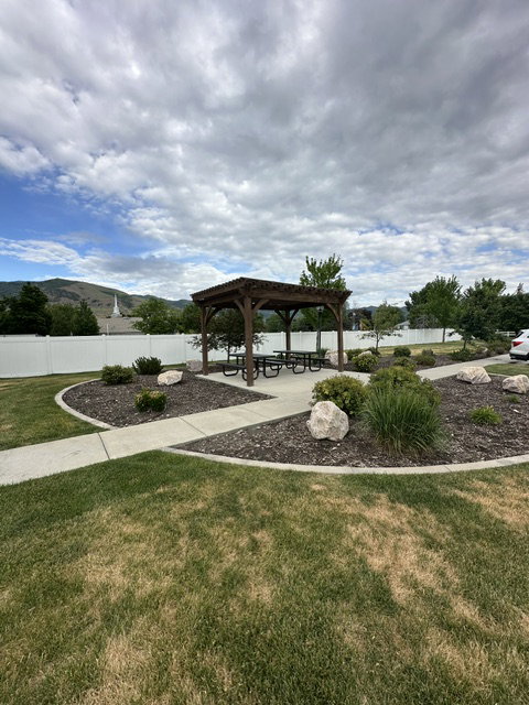 Wood pergola with picnic tables, surrounded by a landscaped area with mature shrubs and decorative rocks