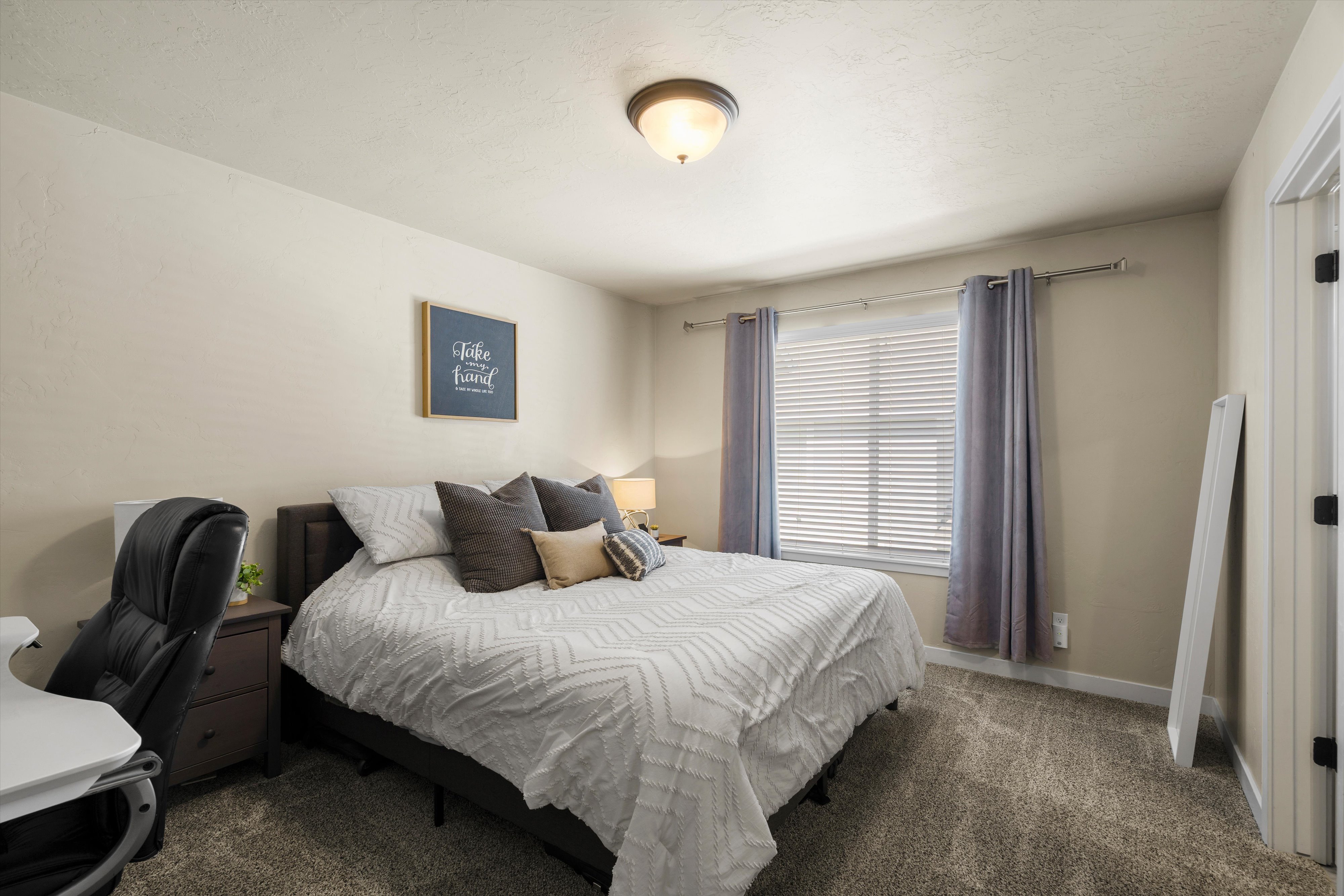 Neutral-toned bedroom featuring wall-to-wall carpeting, a window with blinds and drapes, a flush-mount ceiling light, and a solid core door with dark hardware