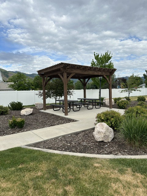 Wood pergola providing a shaded outdoor area, concrete patio, multiple picnic tables, landscaped beds with decorative boulders, and a white vinyl fence